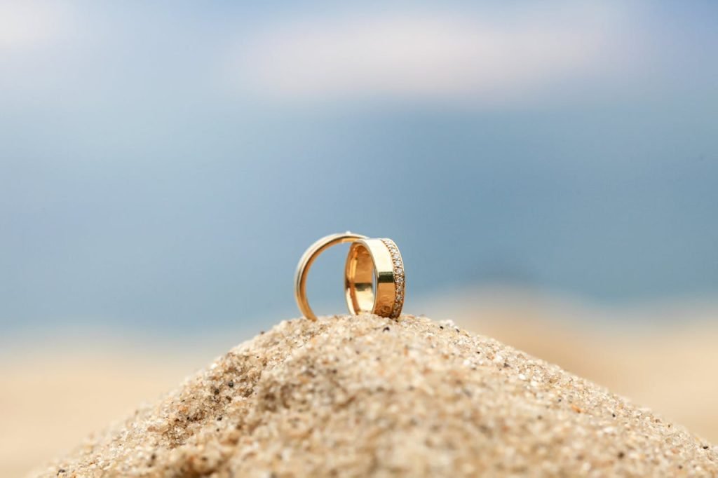 Close-up of elegant wedding rings placed on sand with ocean backdrop, evoking a romantic beach wedding setting.